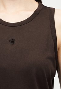 Κοντινό πλάνο ενός ατόμου που φοράει αμάνικο σκούρο καφέ τοπ με μικρό κεντημένο λογότυπο "SB" στο στήθος.