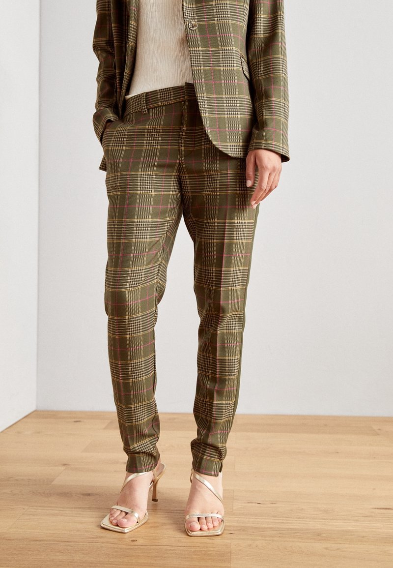 Femme portant un pantalon de costume à carreaux vert olive avec un blazer assorti et des sandales à talons beiges, debout sur un sol en bois.