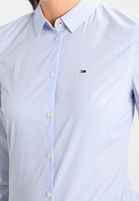 Lyseblå stribet skjorte med knapper og klassisk krave. Har et lille broderet logo på venstre bryst og hvide knapper.