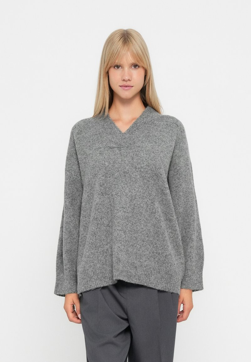 Sweater de lã cinza com decote em V, corte solto e mangas longas. Apresenta uma superfície texturizada e uma bainha assimétrica, oferecendo uma silhueta descontraída.