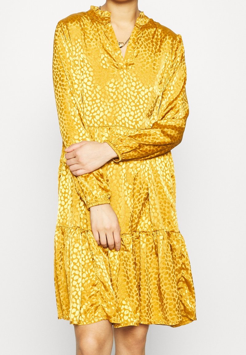 Långärmad, v-ringad klänning i texturerad gul tyg med ett prickigt mönster och en tierad fåll. Mjuk och flödande design.