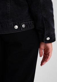 Svart denimjacka med silverfärgade tryckknappar vid ärmslutet, med ett mörkt strukturerat tyg och en rak skärning.