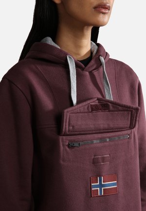 Femme portant un sweat à capuche bordeaux avec des cordons de serrage gris, une poche zippée à l'avant, un rabat avec velcro et un petit patch du drapeau de Norvège.