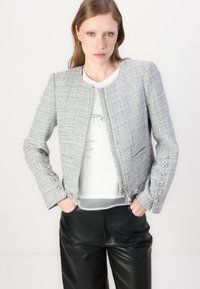Giacca in tweed grigia con design corto, chiusura con zip, bottoni decorativi sulle maniche e orlo sfilacciato. Indossata sopra una t-shirt bianca con grafica.