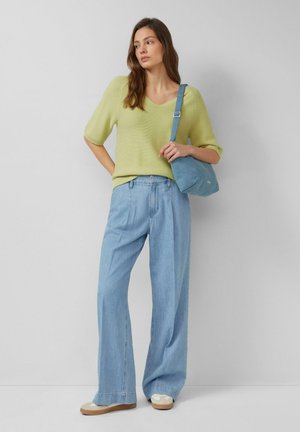 Frau steht vor einer grauen Wand und trägt ein hellgrünes Strickoberteil, lockere hellblaue Jeans, beige Turnschuhe und eine blaue Umhängetasche.