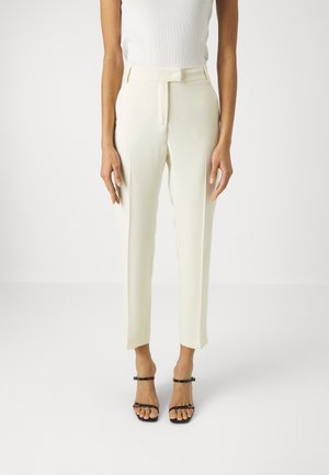 Femme portant un pantalon blanc sur mesure longueur cheville et des sandales noires à talons hauts à brides, debout devant un fond blanc uni.