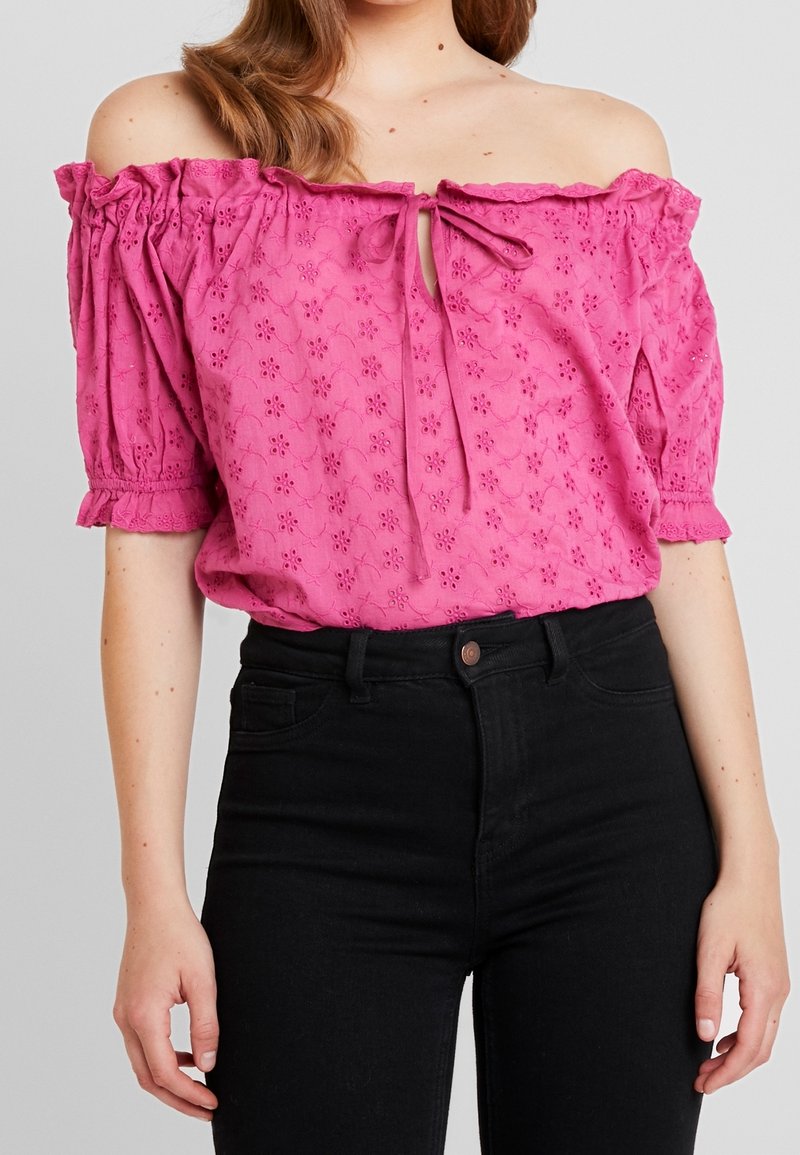 Haut rose décolleté épaules avec broderie florale, détail nœud à l'encolure, et manches courtes bouffantes, associé à un jean noir taille haute.