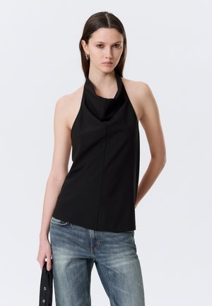 Femme aux cheveux bruns raides portant un top noir sans manches et un jean bleu délavé, tenant un tissu noir dans la main gauche.