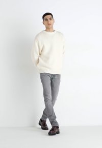 Pull côtelé blanc, jean gris slim-fit et chaussures marron foncé. Le modèle se tient avec les bras relâchés sur les côtés, faisant face à l'avant contre un fond uni.