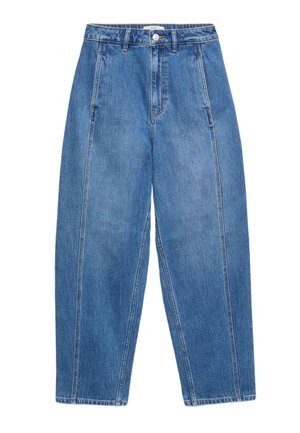 Kvinde med langt krøllet hår iført en sort ærmeløs top, blå jeans og sorte og hvide sneakers, stående med ryggen let vendt.