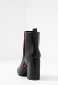 Bottine en cuir noir avec talon bloc, dotée de bandes latérales rouges et bleues et d'une languette arrière, vue de dos sur une surface blanche.