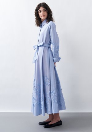 Mujer con un vestido largo de rayas azul claro con bordados florales, un cinturón atado a la cintura y zapatos planos negros, de pie frente a una pared lisa.