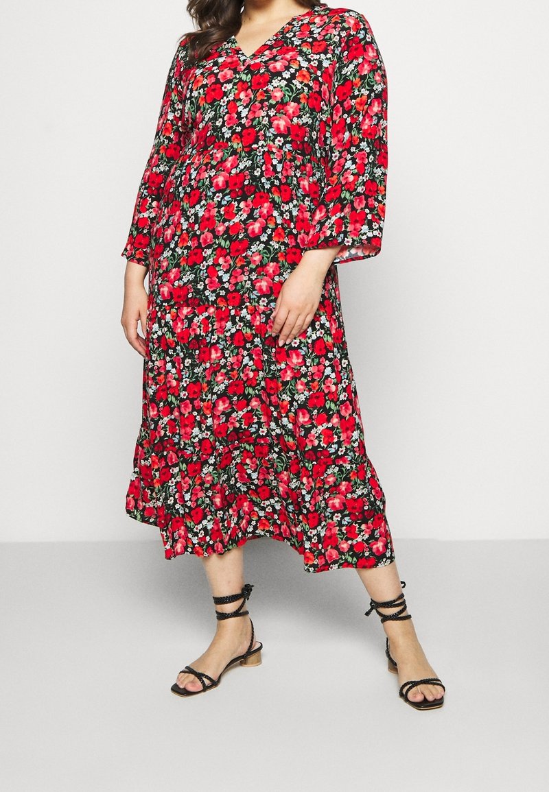 Femme portant une robe à manches longues, longueur mi-mollet, avec un motif floral rouge et des sandales noires à talons bas et brides sur un fond uni.