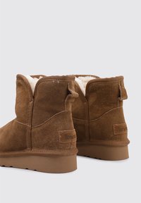 Botines marrones de ante con un suave forro de piel blanca, con suela plana y pequeñas lengüetas en la parte trasera para facilitar su colocación.