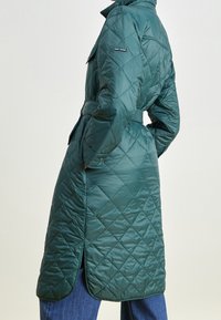 Abrigo acolchado largo verde tipo puffer con cintura ceñida, bolsillos laterales y cuello alto. Presenta un patrón de diamantes y acentos de botones en las mangas.