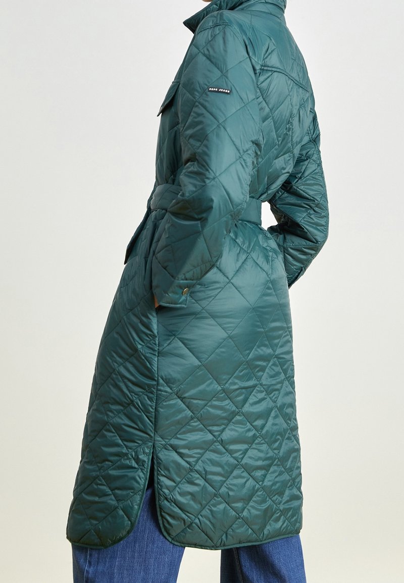 Abrigo acolchado largo verde tipo puffer con cintura ceñida, bolsillos laterales y cuello alto. Presenta un patrón de diamantes y acentos de botones en las mangas.