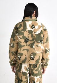 Veste en polaire au motif camouflage, présentant des tons terreux de vert, beige et blanc. Texture douce avec un design col et des poignets élastiques.