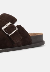 Σκούρο καφέ σουέντ σανδάλι slide με φαρδιές λουρίδες και ασημένια αγκράφα σε μαύρη σόλα, μπροστά σε απλό λευκό φόντο.