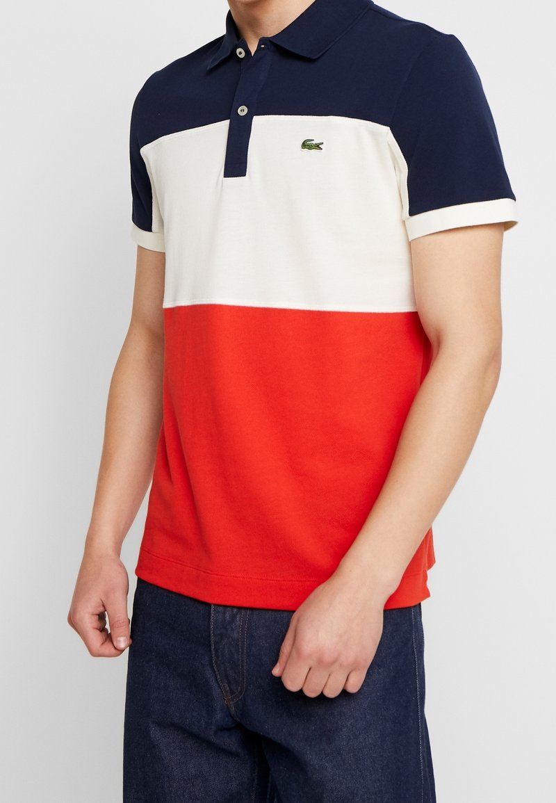 Homme portant un polo color-block bleu marine, blanc et rouge et un jean en denim bleu foncé, sans détails visibles en arrière-plan.