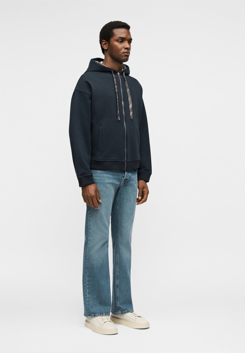 Homme portant un sweat à capuche noir zippé avec des cordons à motifs, un jean bleu clair et des baskets blanches, debout devant un fond uni.