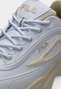 Λευκό και μπεζ παπούτσι FILA με ανοιχτό μπλε κορδόνια, εμφανίζοντας κεντημένο λογότυπο στη γλώσσα και στην πλευρά κοντά στον επενδεδυμένο γιακά.