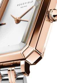 Reloj Rosefield con una esfera blanca estampada, caja de oro rosa y pulsera de metal plateado. Presenta una forma hexagonal y una corona texturizada.