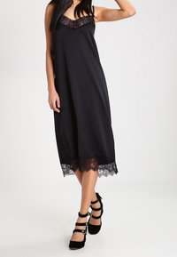 Musta ilmanhiha dressi, jossa on pitsireunat kaula-aukossa ja helmassa, sileää kangasta, löysä siluetti ja elegantti laskostus.