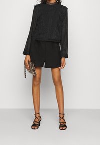 Musta, tekstuuriga pikakäiseline pluus rippuvate volangidega, koos mustade lühikeste pükstega ja rihmadega mustade sandaalidega, hoides leopardi trükkimisega kotti.