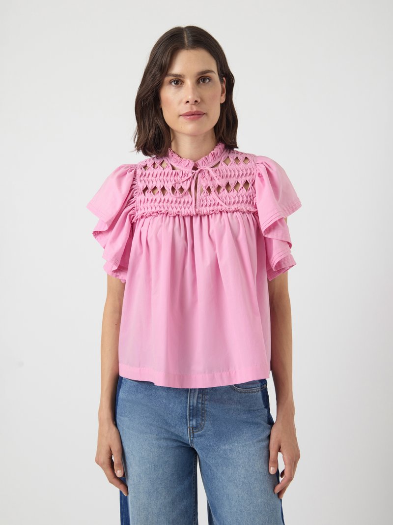 Femme portant une blouse rose ample avec des manches froncées et un haut à smocks et découpes, associée à un jean en denim bleu.