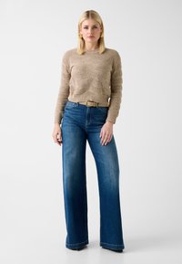Pull beige en tricot avec motif texturé, assorti à un jean bleu taille haute à jambes larges et une ceinture beige, sur un fond blanc.