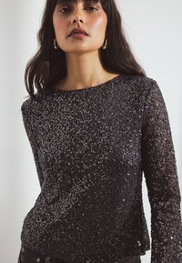Črn dolg rokav majica iz sequins tkanine, ki ima okroglo izrez in teksturirano površino z bleščečim učinkom.