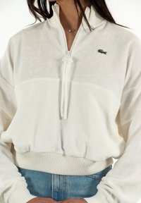 Sudadera cremosa con cremallera y un acabado texturizado. Presenta una cintura y puños acanalados, con un pequeño logo verde en el pecho.