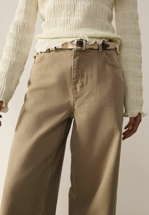 Persoană care poartă pantaloni bej cu talie înaltă și croială largă, cu o centură de piele maro uzată și un top crem texturat cu mâneci lungi.