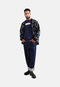 Cárdigan negro con patrones florales azules, camiseta navy con logo, jeans azules holgados y botas negras de tobillo con suelas texturizadas.