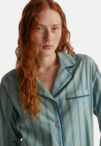 Pruhovaná bavlněná pyžamová košile v světle modré a bílé barvě s náprsní kapsou, zapínání na knoflíky a kontrastním tmavě modrým lemováním kolem límce.