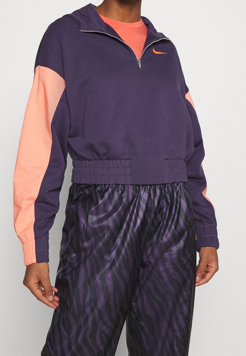 Fialová mikina na zip s korálovými rukávy, elastickým pasem a malým oranžovým logem. Kombinováno s volnými kalhotami v fialovo-černém zebrovém vzoru.