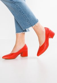 Füße in leuchtend roten Blockabsatzschuhen, kombiniert mit ausgefransten blauen Jeans, heben einen Fuß vom weißen Boden.