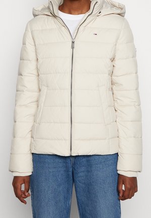 Person iført en beige quiltet jakke med hætte og lynlås, kombineret med blå jeans, foran en ensfarvet hvid baggrund.