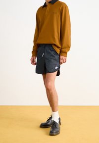 Brun långärmad polo med svart krage, matchad med svarta mönstrade shorts med en vit logotyp, och svarta skor med vita strumpor.