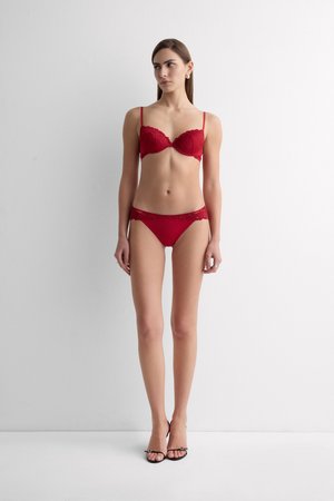 Femme debout contre un mur blanc portant un soutien-gorge en dentelle rouge et une culotte assortie avec des sandales à talons noires, regardant légèrement à gauche.