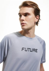 Camiseta deportiva de color gris claro con el texto negro "FUTURE". Tiene manga corta, cuello redondo y una tela texturizada.