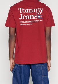 T-shirt en coton rouge avec un texte blanc portant "Tommy Jeans" et divers noms de villes. Col rond et manches courtes, texture lisse.