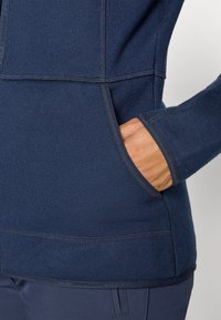 Person som har på seg en marineblå strikkegenser med den ene hånden delvis inne i en frontlomme, kombinert med mørkeblå bukser.