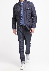 Laivastonsininen tikattu takki, jossa pystykaulus ja kaksi rintataskua, yhdistetty vaaleansiniseen kauluspaitaan ja tummiin farkkuihin. Valkoiset lenkkarit.