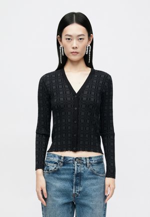 Cardigan negru texturat cu decolteu în V, mâneci lungi și nasturi frontali. Design cropped cu un model geometric. Purtați peste blugi bleu deschis.