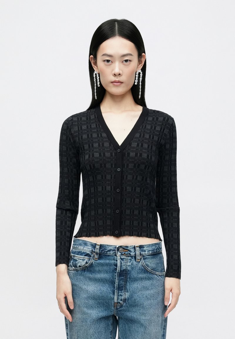 Cardigan negru texturat cu decolteu în V, mâneci lungi și nasturi frontali. Design cropped cu un model geometric. Purtați peste blugi bleu deschis.