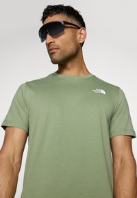 Camiseta de algodón verde con mangas cortas y un logo blanco en el hombro izquierdo, combinada con unas gafas de sol oscuras y atrevidas con un diseño deportivo.
