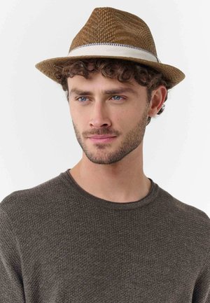 Junger Mann mit lockigem Haar und hellem Bart, der einen braunen, strukturierten Pullover und einen braunen, gewebten Fedora-Hut mit weißem Band trägt.