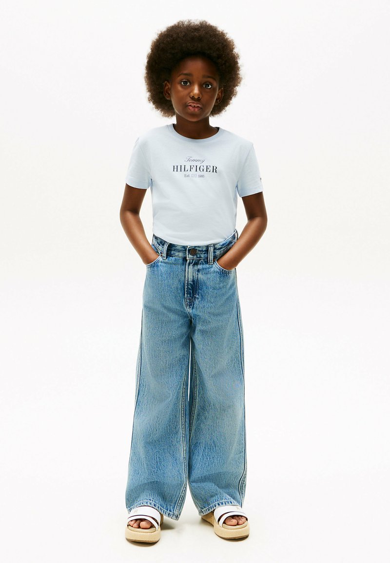 Una camiseta de color azul claro con el logotipo negro "Tommy HILFIGER", combinada con unos vaqueros de corte holgado y de lavado claro. Sandalias de plataforma blancas completan el look.