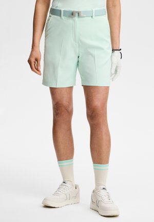 Mand iført lette mintgrønne skræddersyede shorts, matchende bælte, hvid golfhandske, hvide sneakers og hvide sokker med blå striber, stående.